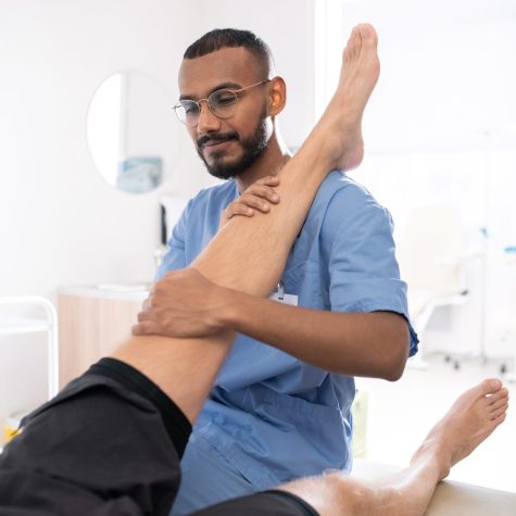 Young contemporary mixed-race professional holding sick leg of his patient while massaging it in hospital